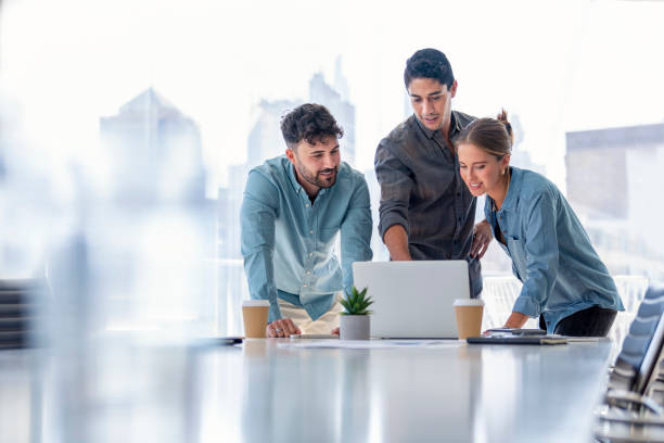 Business team working on a laptop computer. Three people are wearing  casual clothing. They are standing in a board room. Multi ethnic group with Caucasian and Latino men and women